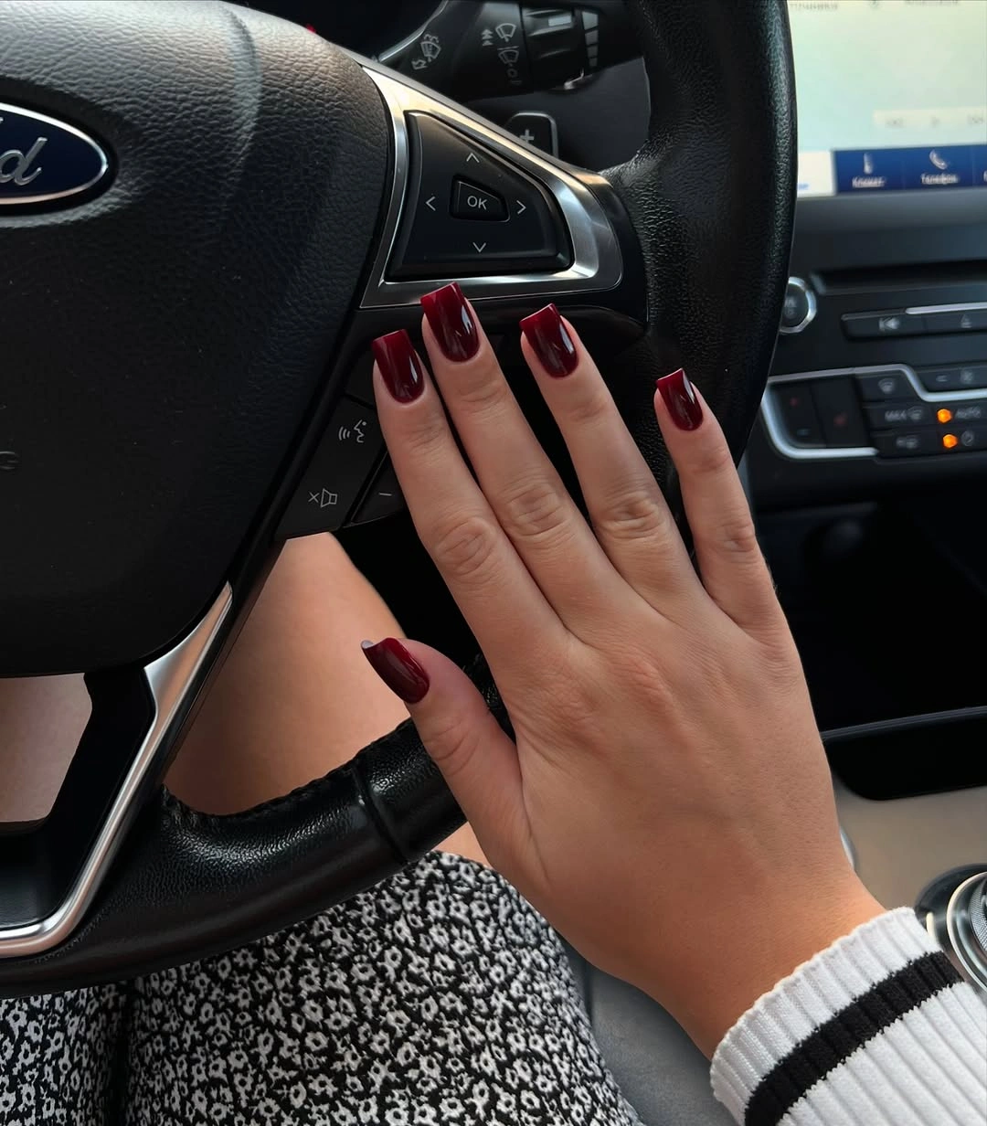 Short square burgundy nails near a car steering wheel