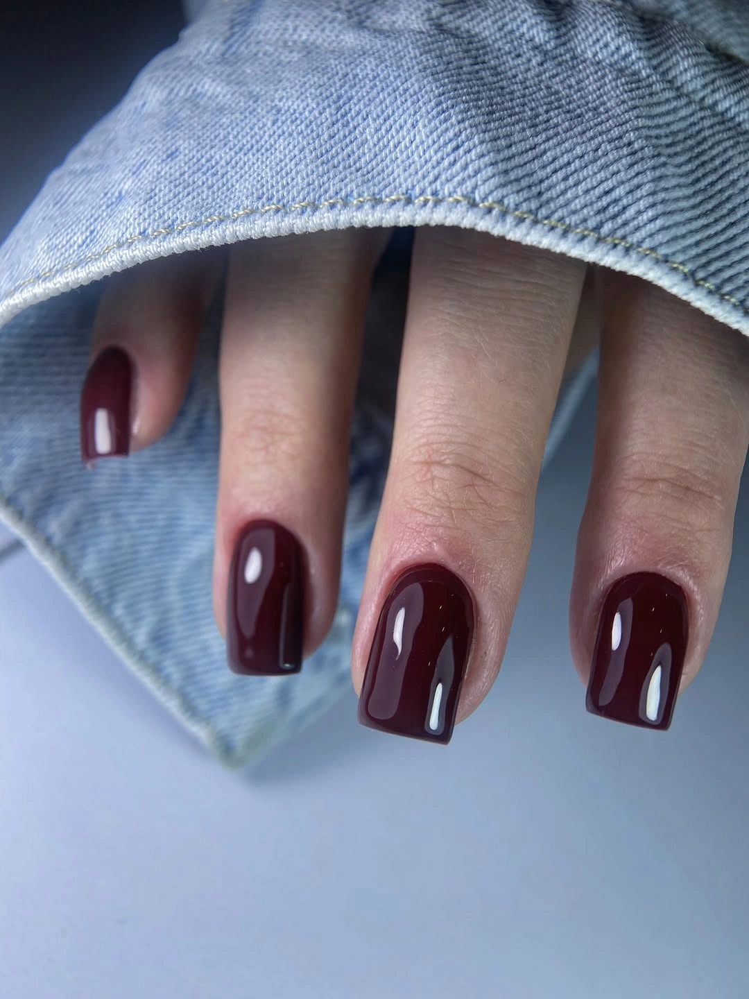 Short square burgundy nails styled against blue denim