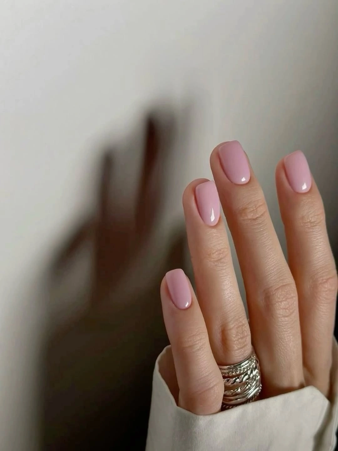 Short oval nails in a soft glossy pink color against a warm wall