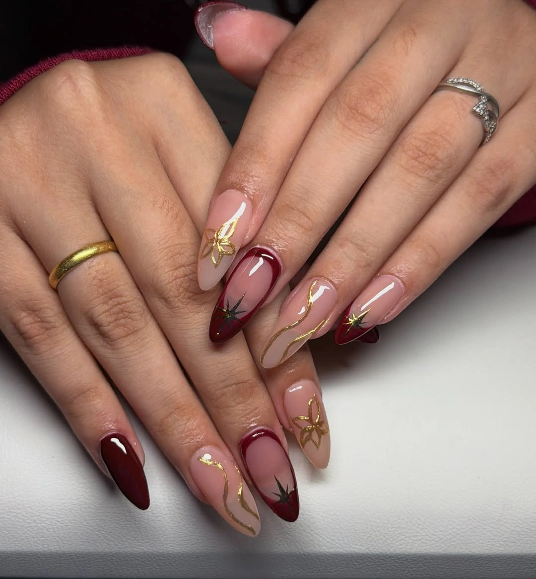 Nude almond nails with burgundy curved tips and small gold botanical details