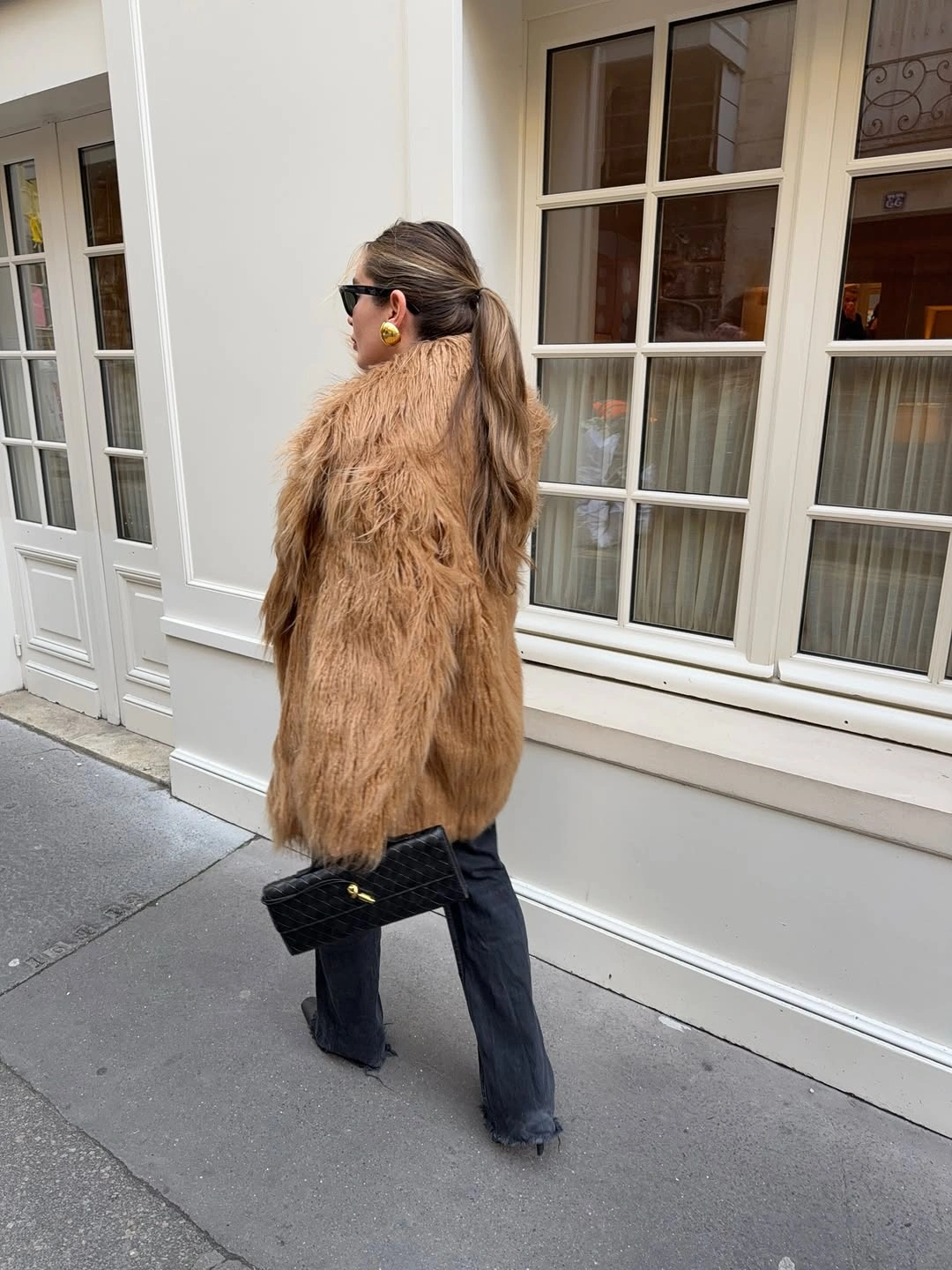 Long tan textured coat with wide dark jeans and black clutch