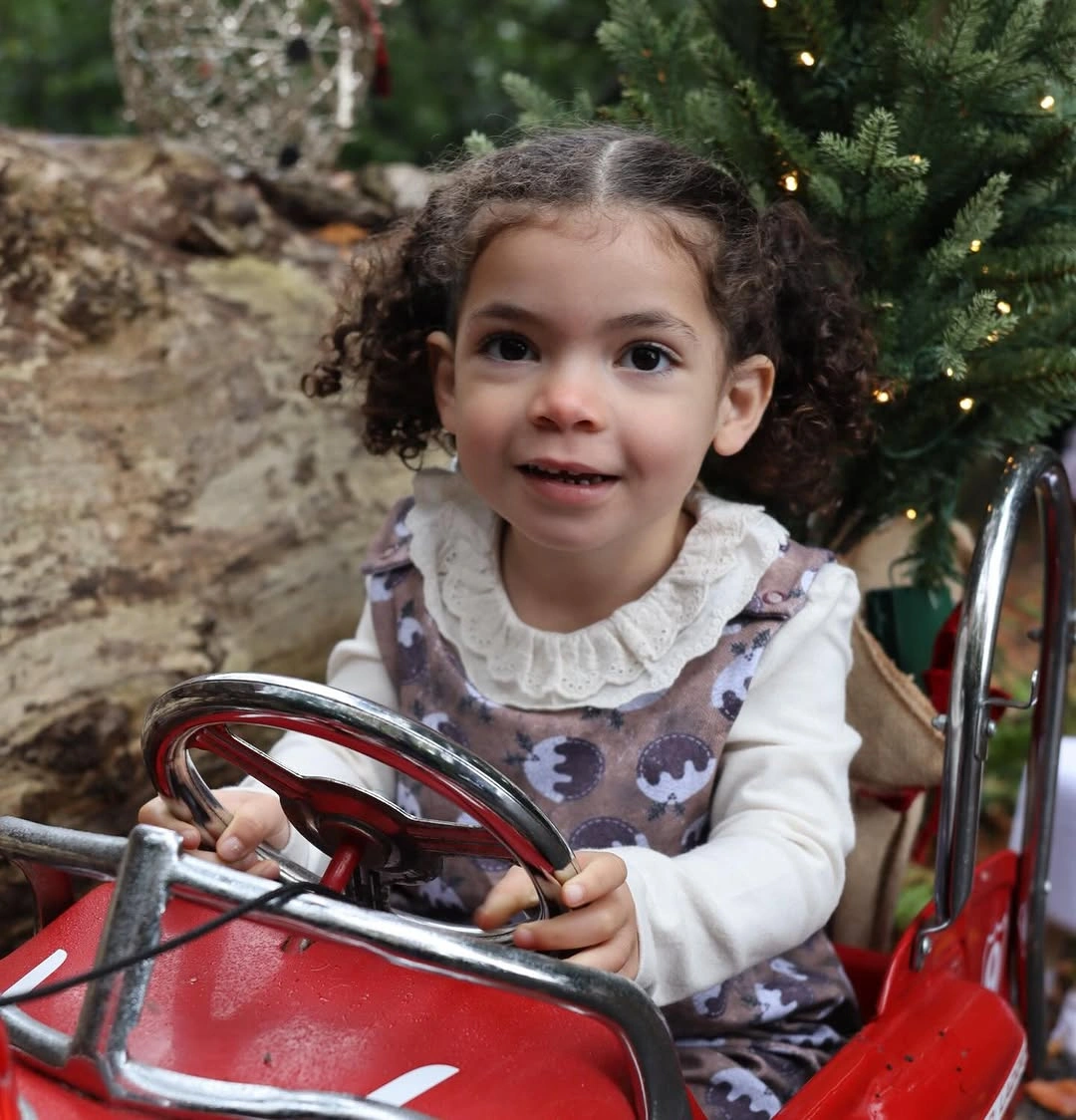 Child wears patterned pinafore dress over white ruffled top