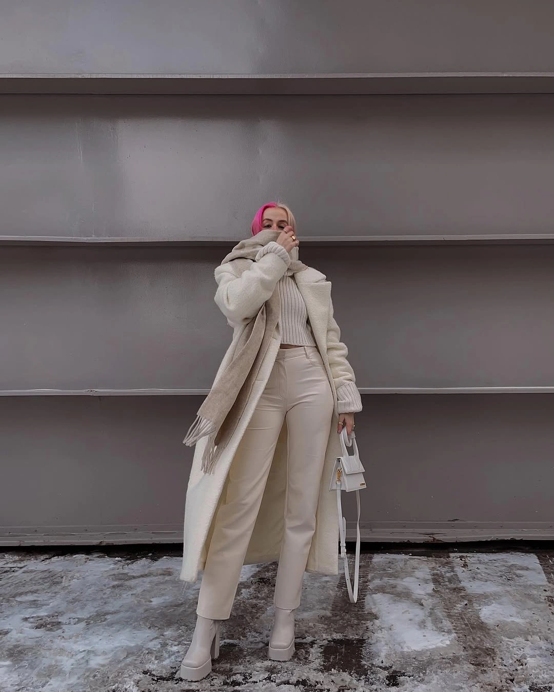 Long light coat with cream knitwear, white trousers, scarf, and small bag in snow