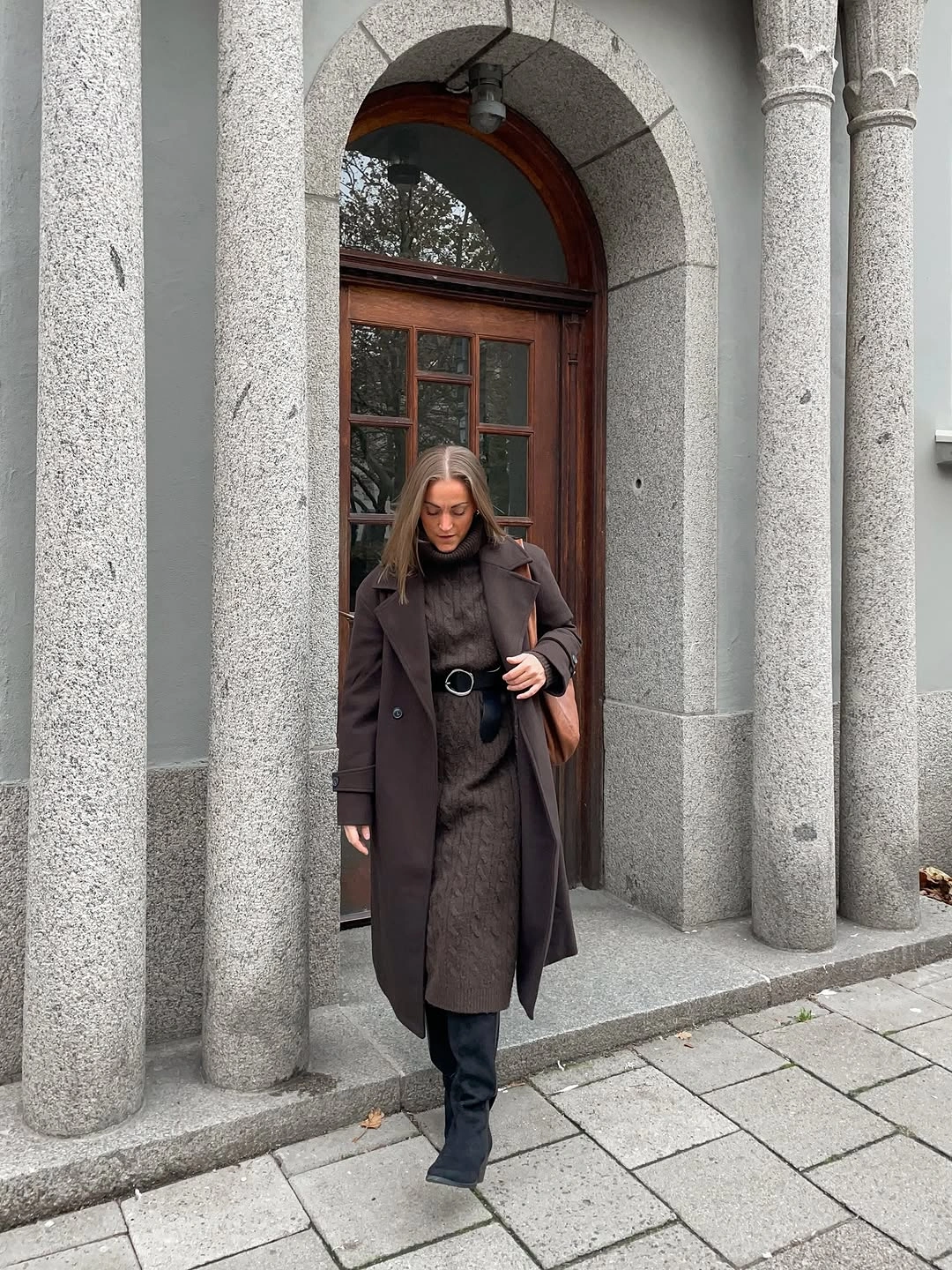Dark brown belted long coat with dark boots and structured bag