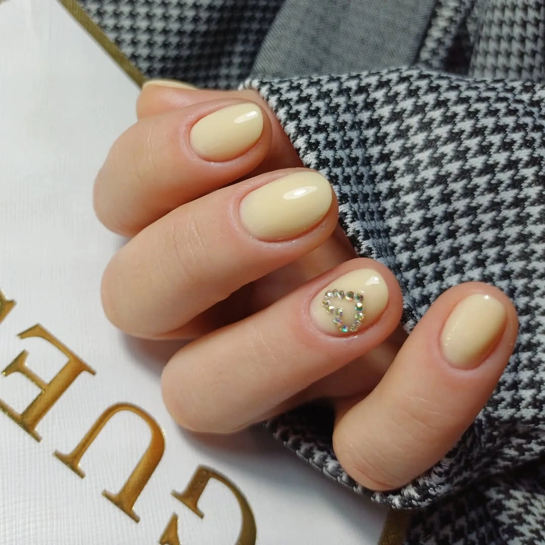 Short creamy yellow nails with silver-toned heart accent