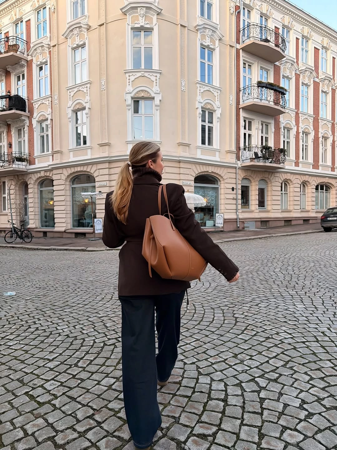 Dark coat, wide dark trousers, tan tote, cobblestone street