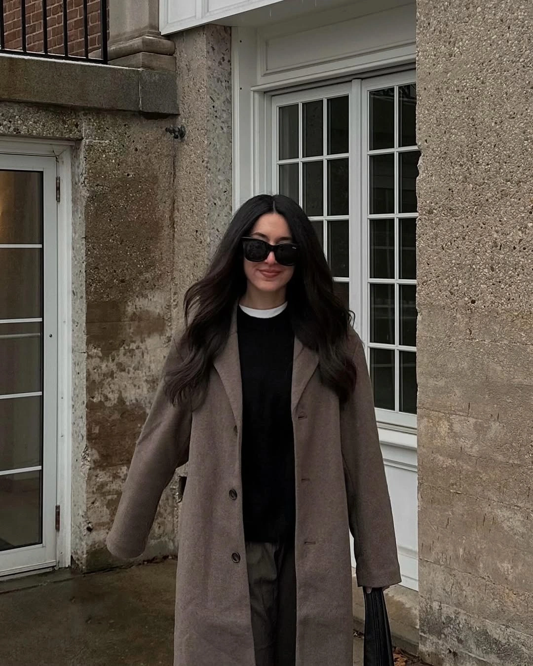 Brown coat over black top and trousers with black sunglasses and handbag