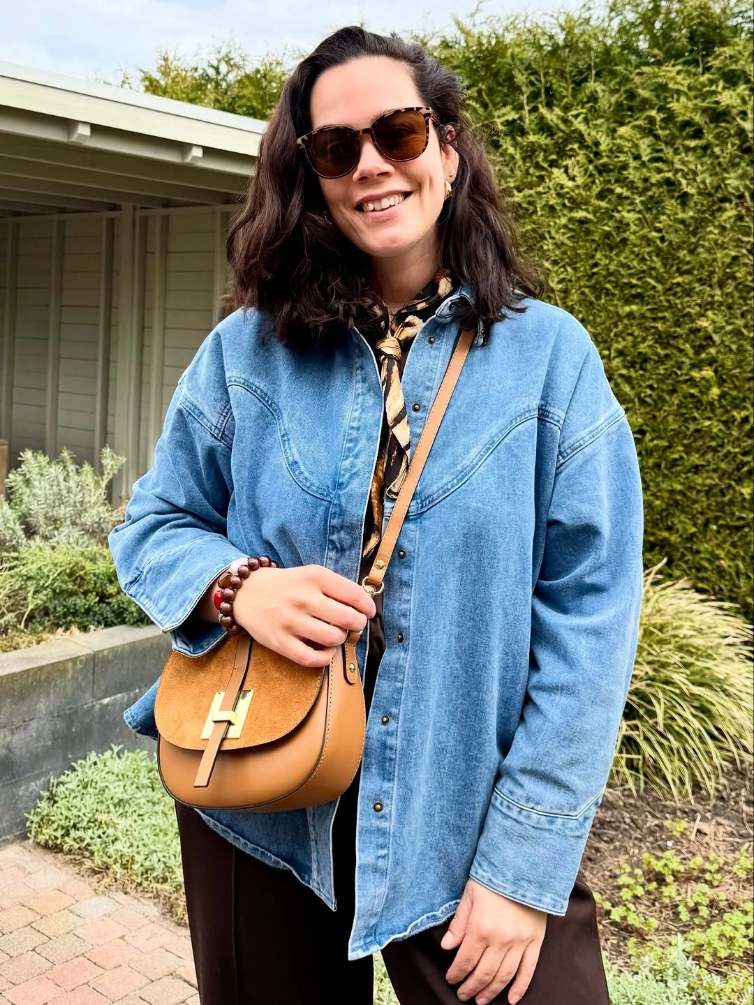Woman in taupe jacket, dark wide jeans, cap, and crossbody bag