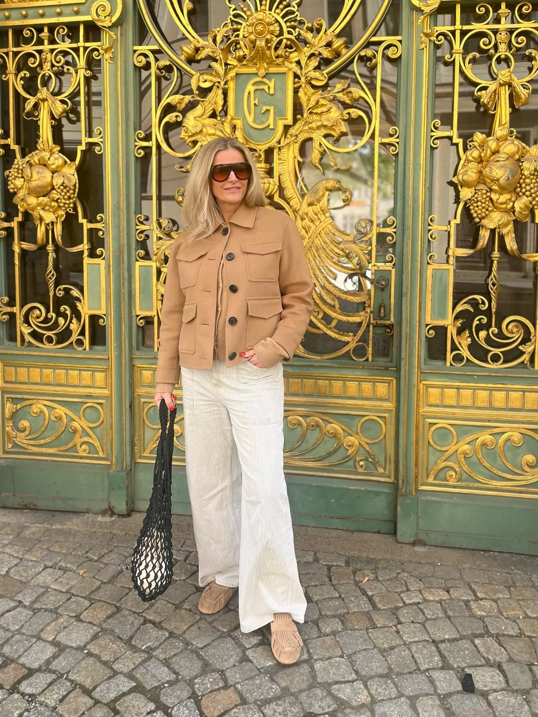 Woman in camel jacket, white wide trousers and net bag