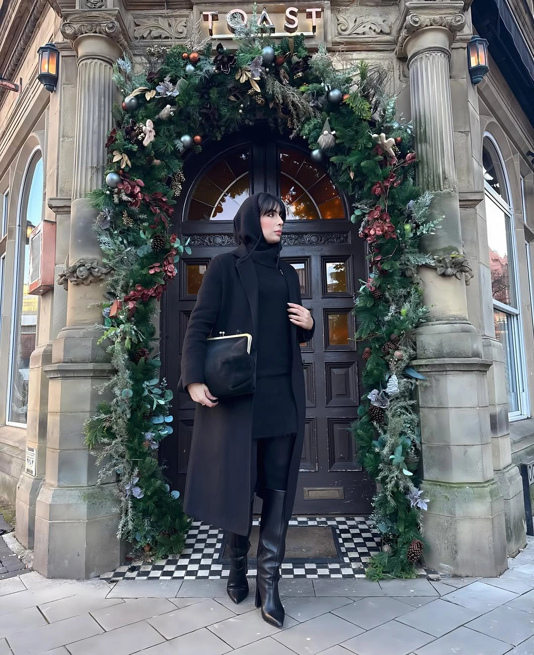 Long black coat with high-neck layer, tall boots, and clutch