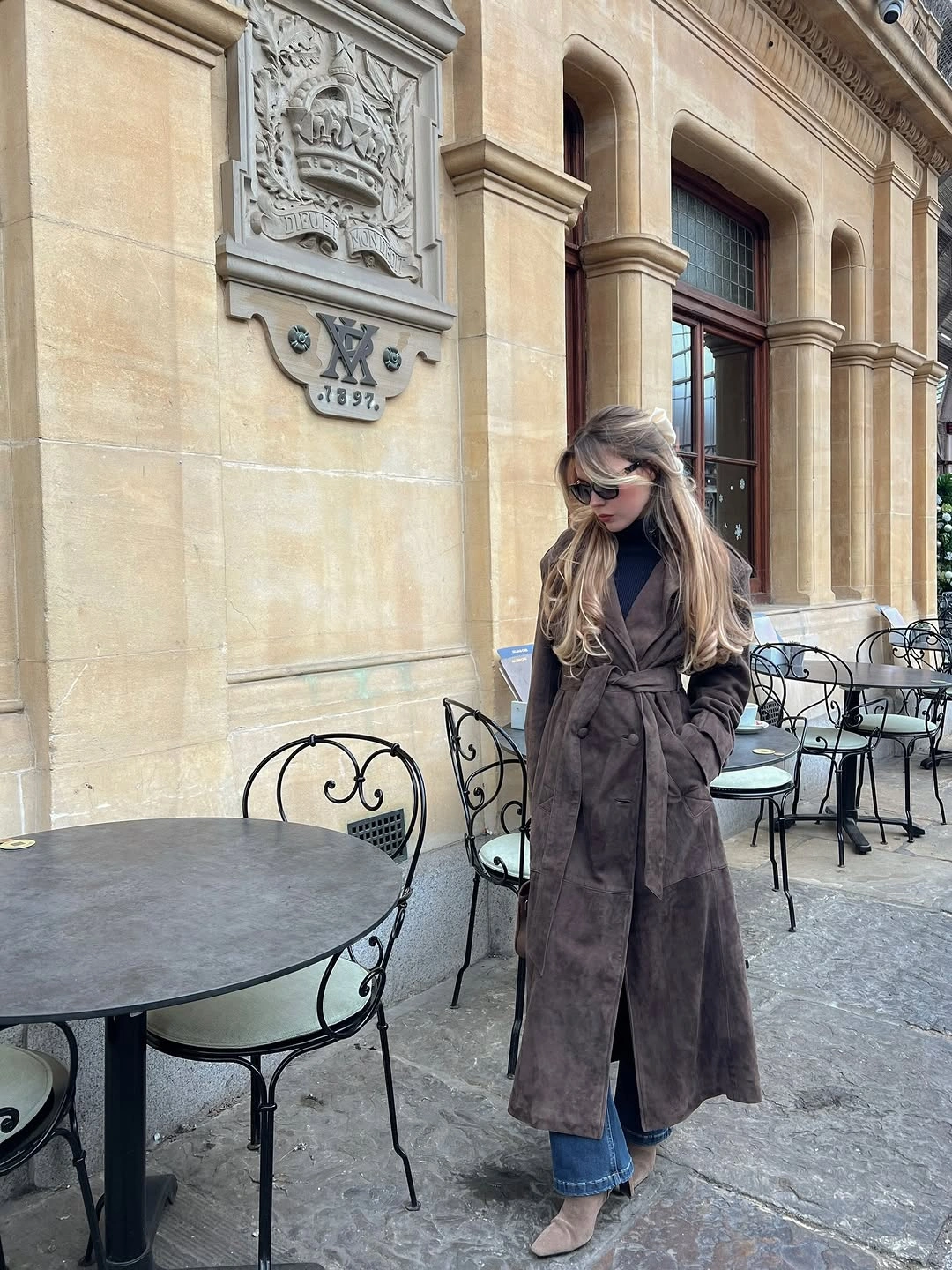 Long brown belted coat with black top, jeans, and beige shoes