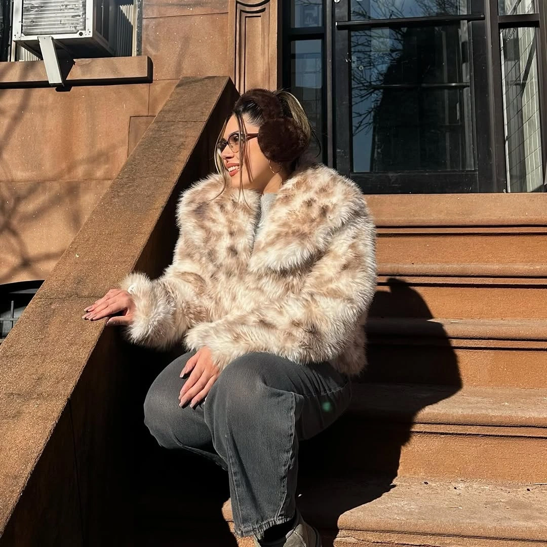Woman in fur-looking jacket, charcoal jeans, earmuffs, and sunglasses