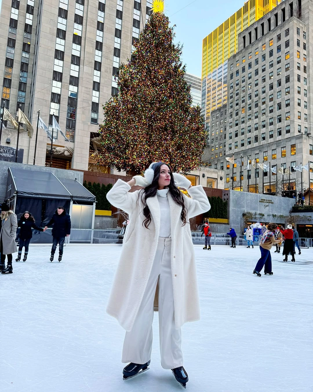 Woman in long white coat, wide-leg trousers, earmuffs, and skates
