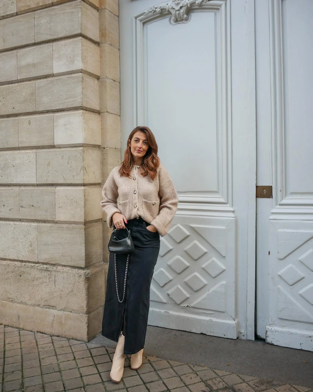 Beige cardigan with dark front-slit skirt, pale boots, and black handbag