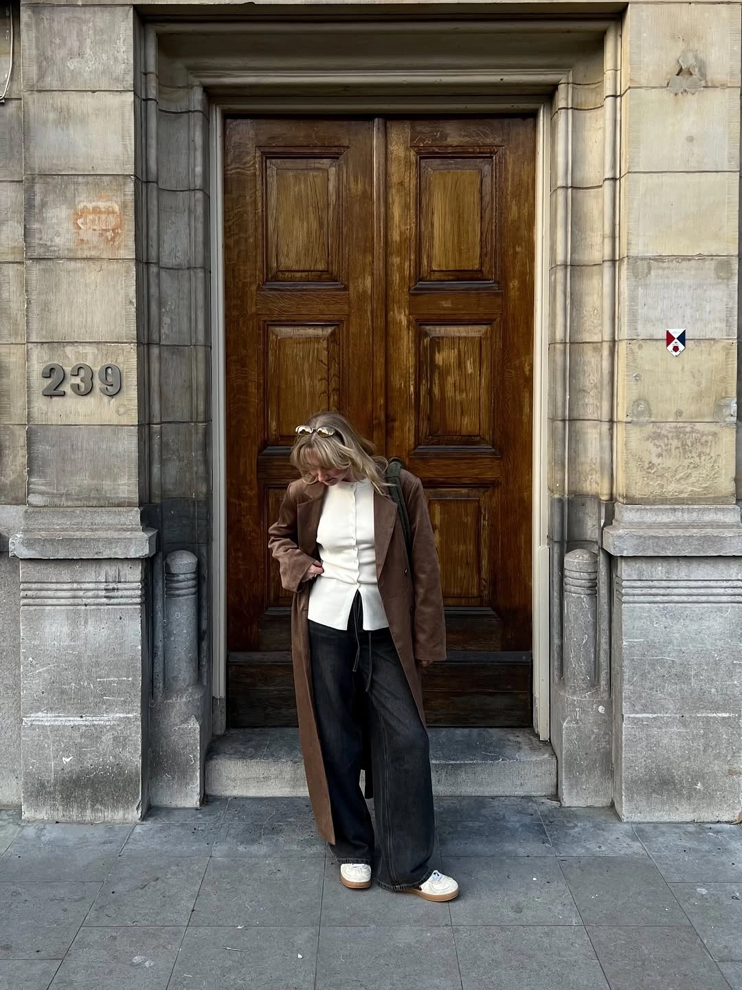Woman in brown long coat, white fitted top, and dark wide-leg bottoms