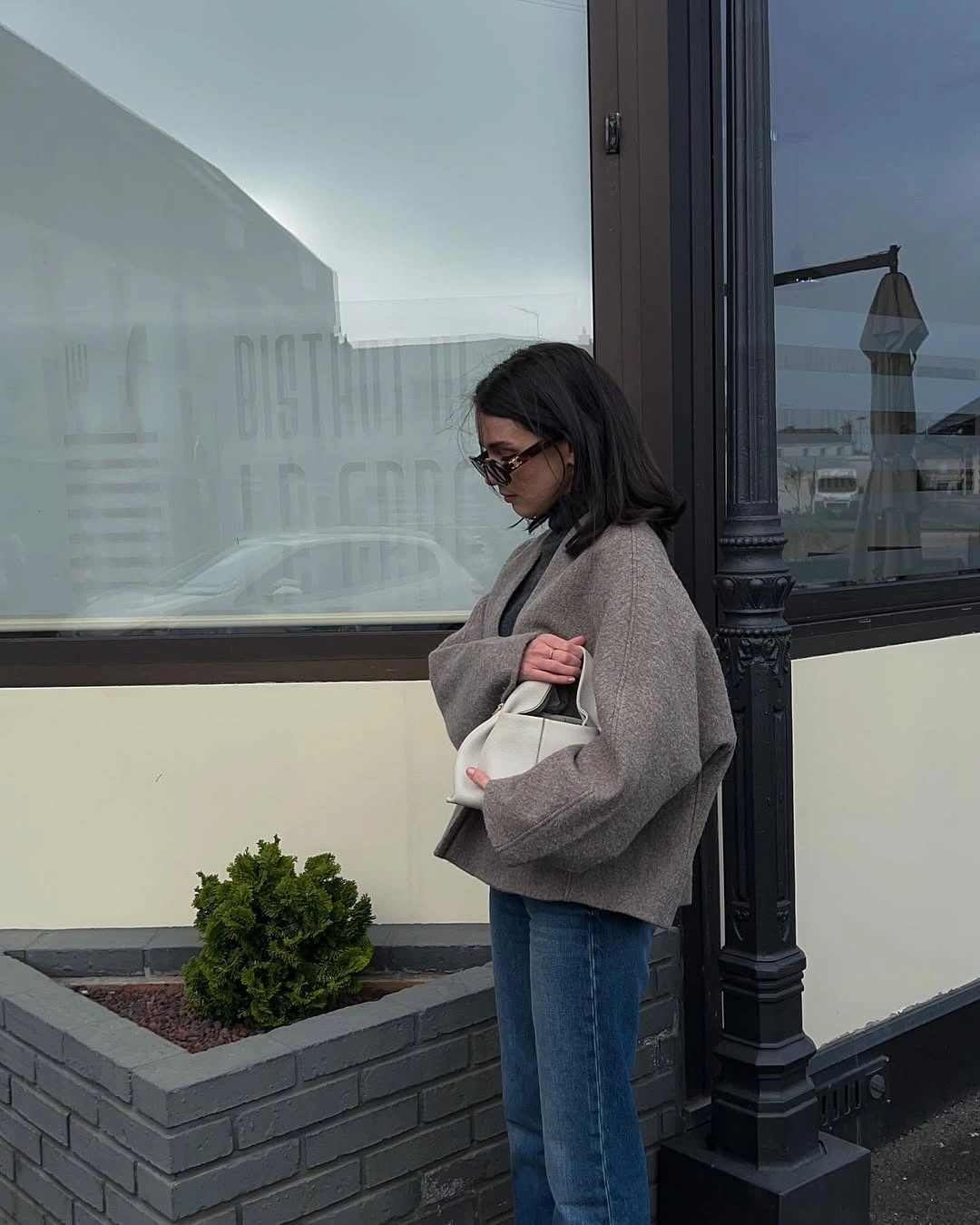 Woman in taupe sweater, straight blue jeans, and white handbag