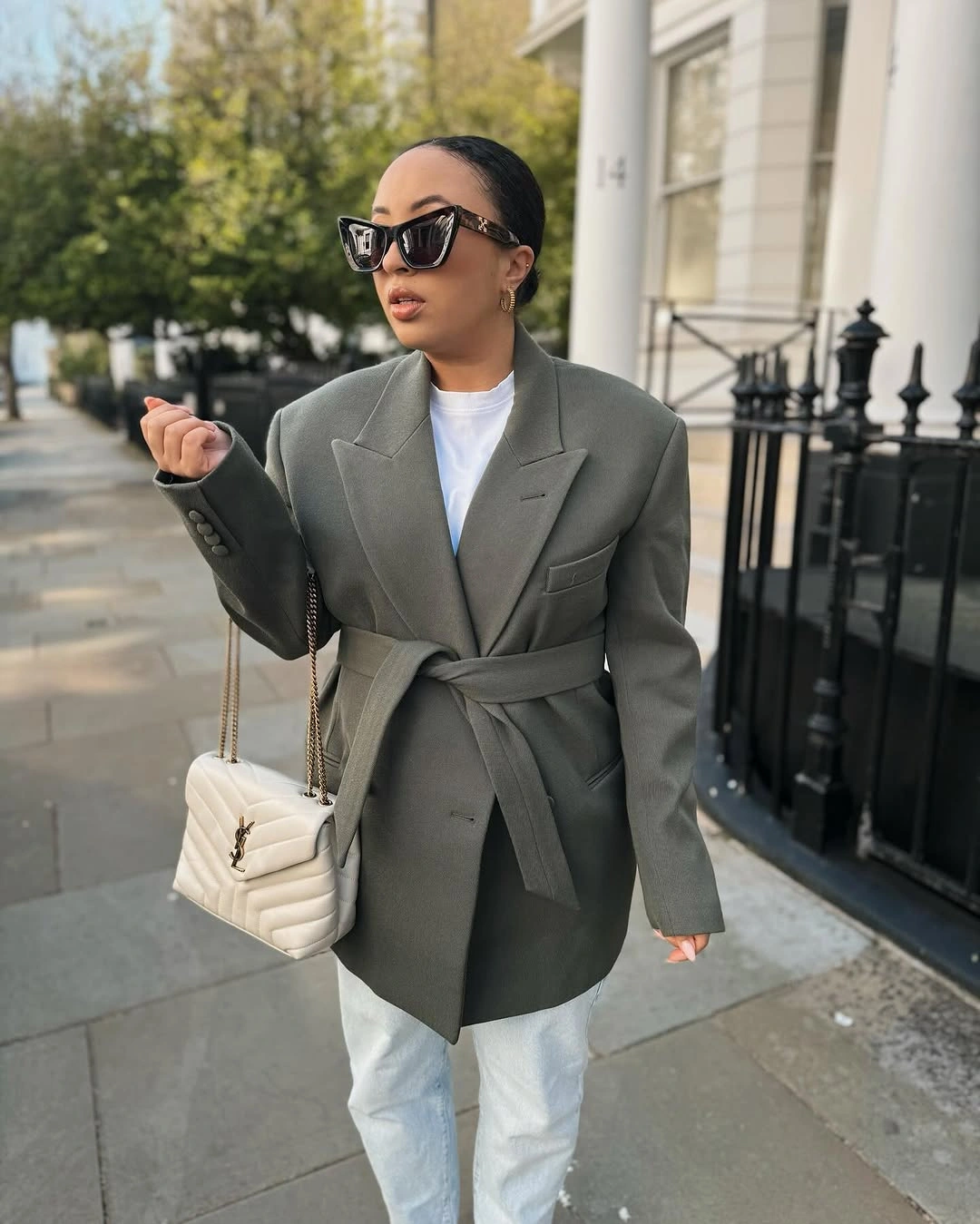 Woman wearing belted gray-green blazer, pale jeans, and cream bag