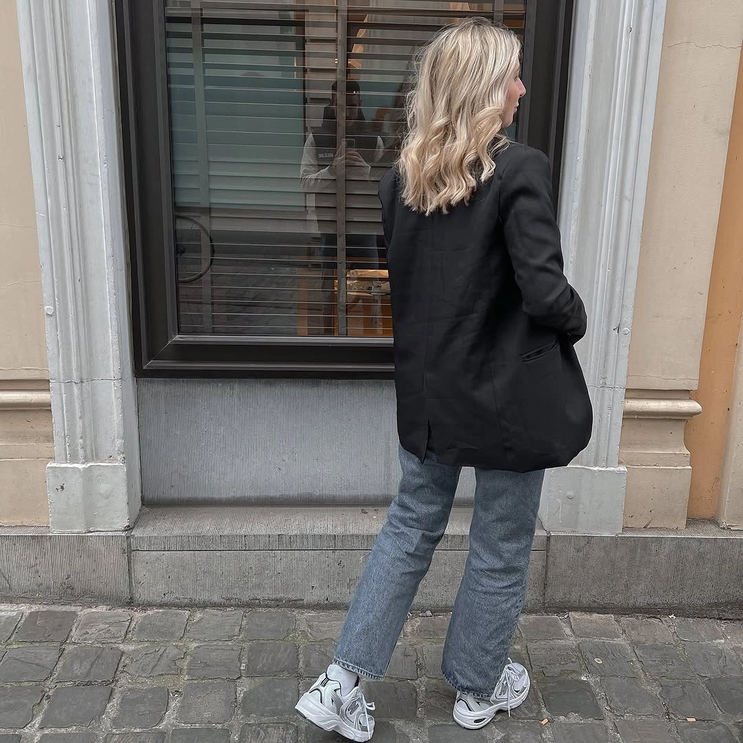 Woman wearing black blazer, gray-blue jeans, and chunky gray sneakers