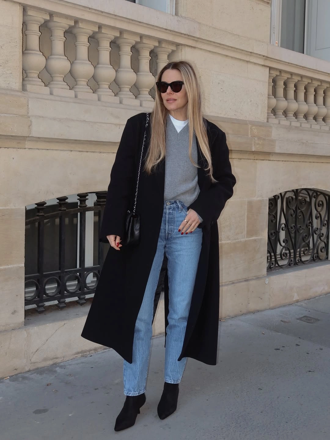 Long black coat over gray V-neck layer, blue jeans, and pointed boots