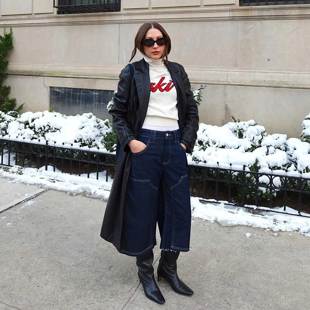 Woman wearing long dark coat, white graphic top, jeans, and black boots