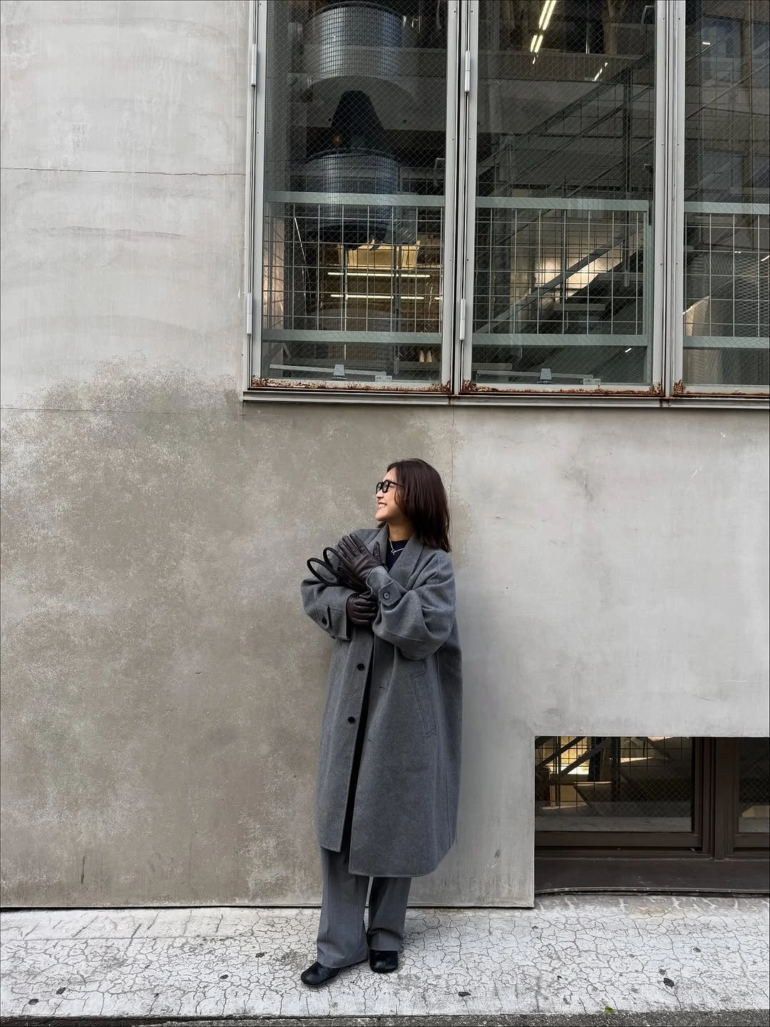 Woman wearing long charcoal coat, dark trousers, sunglasses, and black shoes