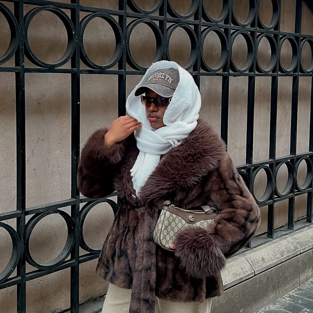 Woman in fluffy brown jacket, white scarf, gray cap, and patterned handbag