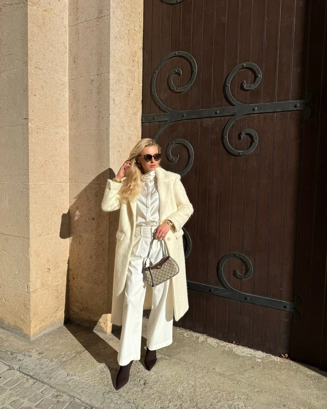 Woman wearing cream coat, pale trousers, dark shoes, and patterned handbag