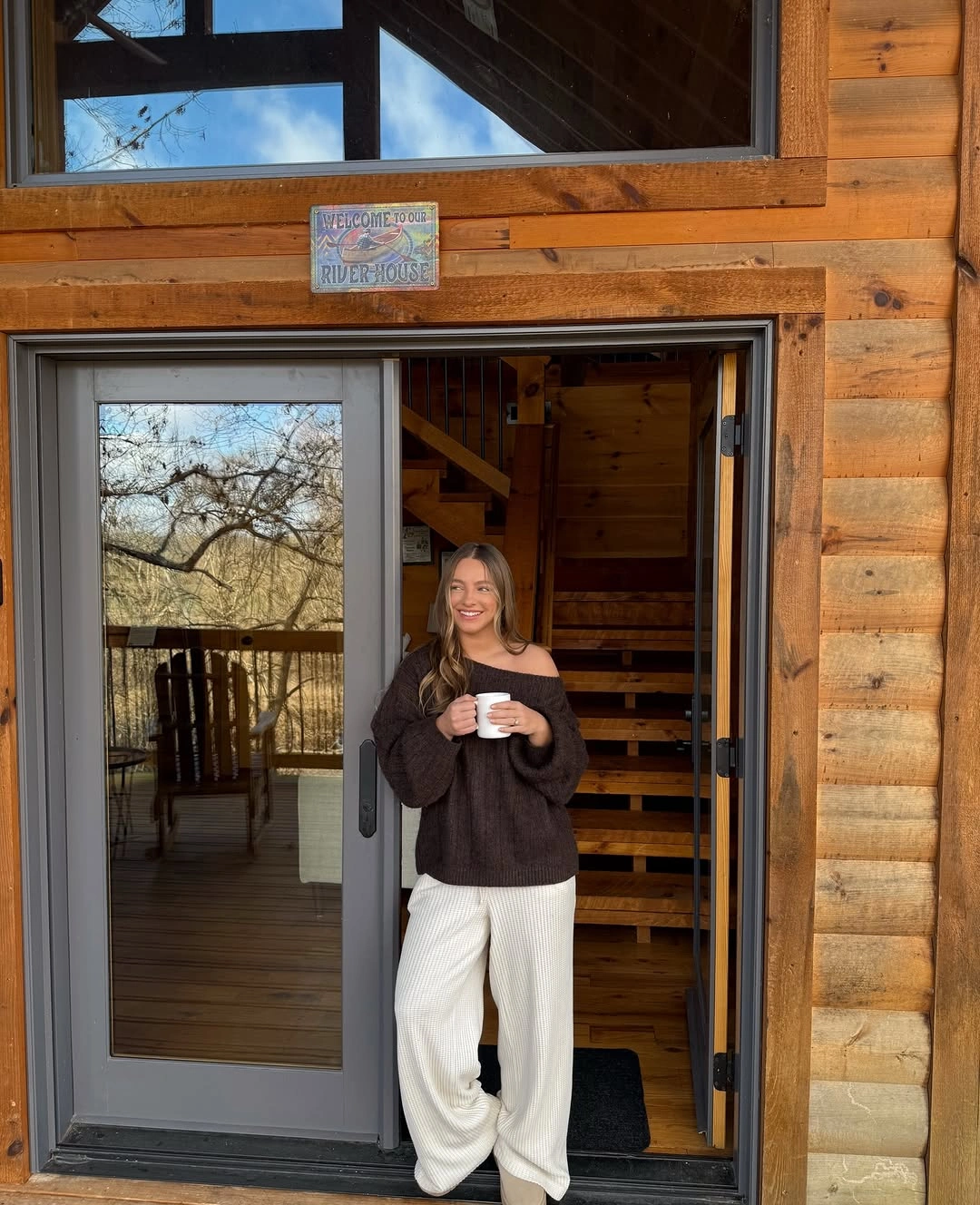 Woman in dark brown sweater and wide cream trousers holding a cup