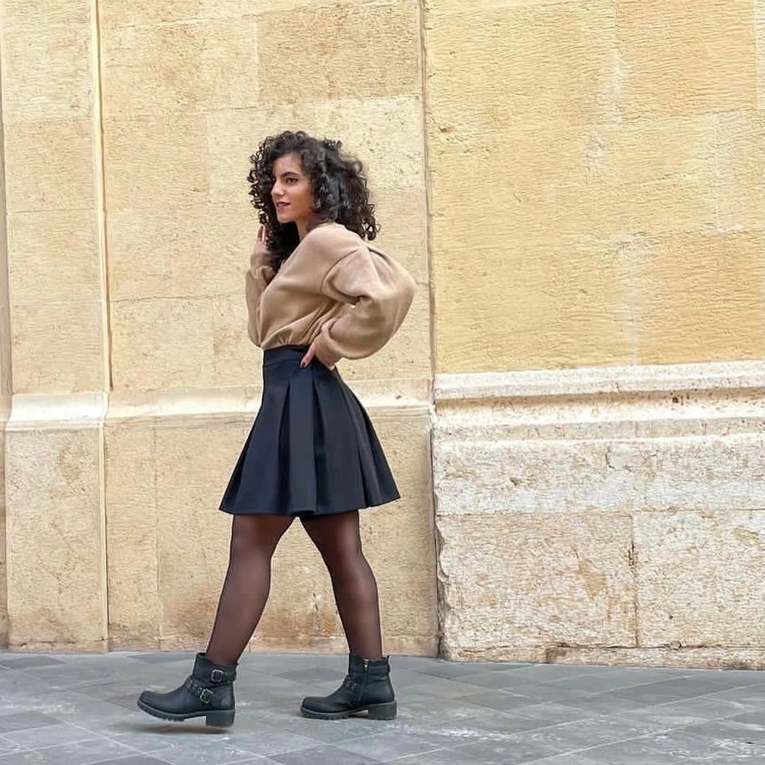 Beige oversized sweater, black pleated mini skirt, sheer tights, and lace-up boots