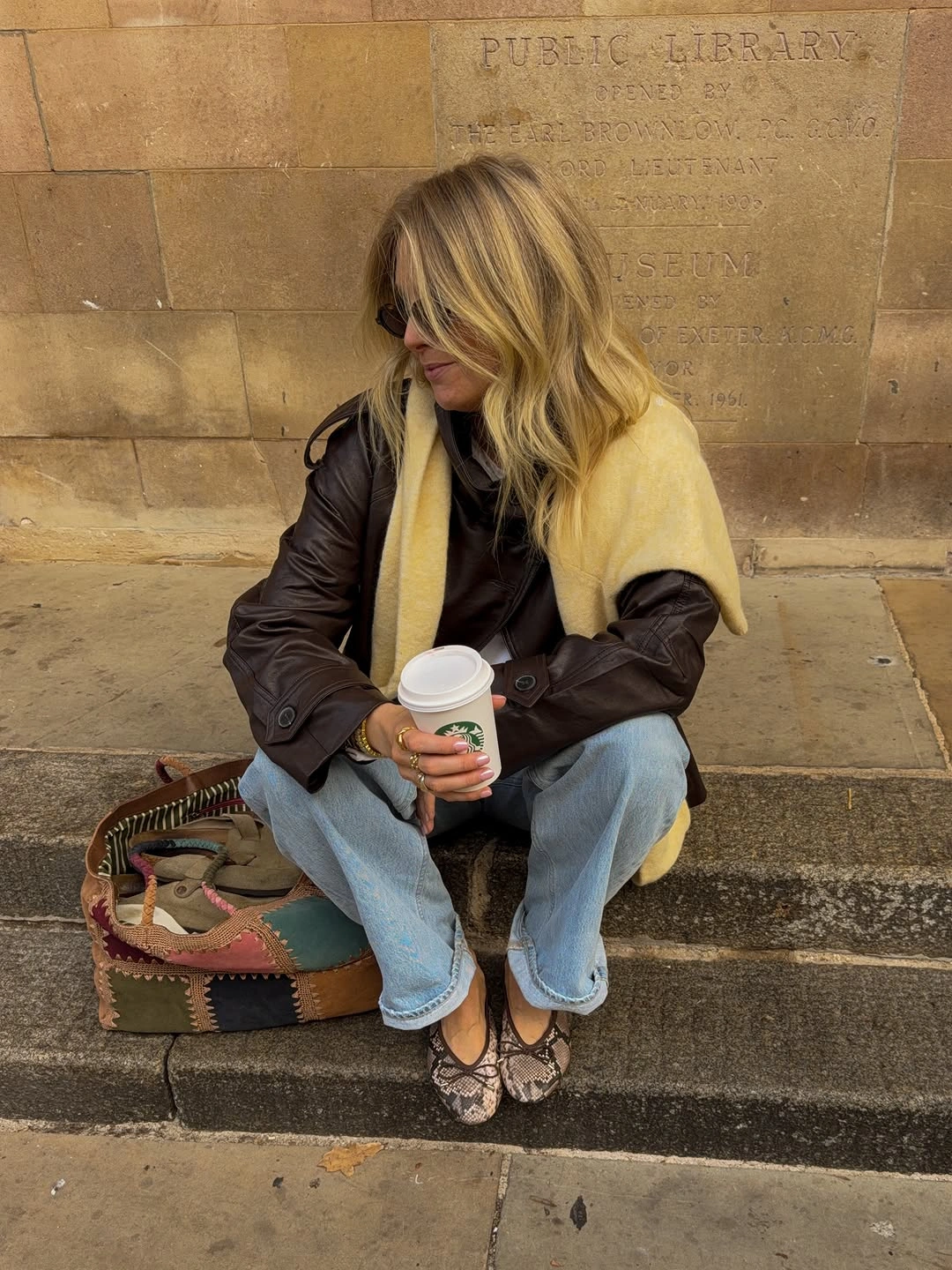Brown leather-look jacket pale yellow scarf blue jeans and patterned flats